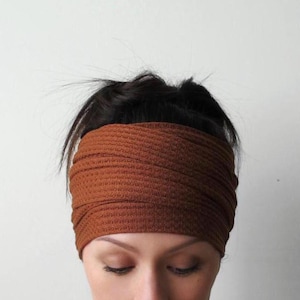 Op de afbeelding: Een brede, roestkleurige hoofdband van een textielstof. De hoofdband is om het hoofd gewikkeld en bedekt het voorhoofd en de oren. De achtergrond is een effen witte muur.