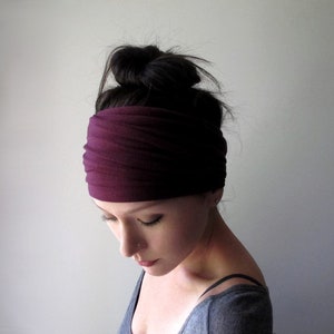 Op de afbeelding: Een brede, effen donkerpruimkleurige stoffen hoofdband met een geplooide, gerimpelde textuur. De hoofdband is horizontaal over het voorhoofd geplaatst, met een zachte, matte afwerking en een flexibele, rekbare constructie die om het hoofd zit.