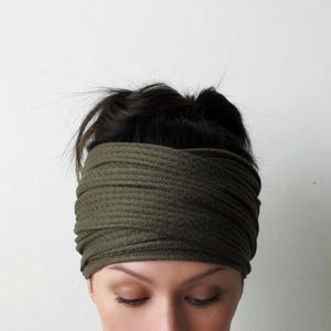 May include: An olive green headband featuring a textured waffle knit fabric. The accessory is designed with a wide, overlapping crisscross pattern at the front, creating a layered appearance that covers the forehead and sits securely around the head.