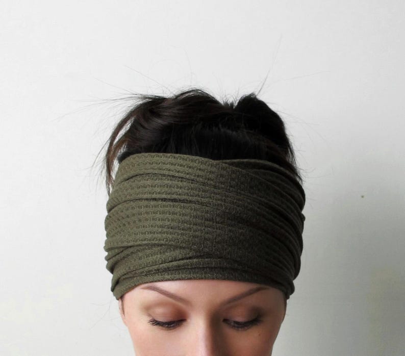 May include: An olive green headband featuring a textured waffle knit fabric. The accessory is designed with a wide, overlapping crisscross pattern at the front, creating a layered appearance that covers the forehead and sits securely around the head.