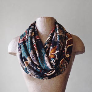 May include: An infinity scarf featuring a black base with a multi-colored paisley pattern in shades of teal, mustard yellow, red, and cream. The fabric is draped in multiple loops around a mannequin neck, displaying intricate floral and teardrop motifs.