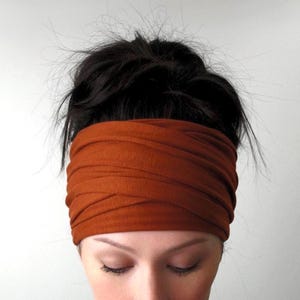 May include: A rust-colored headband made of soft fabric. The wide band wraps around the head, covering the forehead and ears. The headband is worn by a person with dark hair pulled up in a bun.
