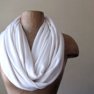 May include: A white infinity scarf made of soft fabric. The scarf is draped over a mannequin's neck and shoulders.