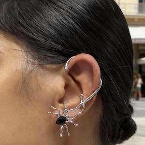 May include: A silver ear cuff featuring a spider design with a black gemstone at its center. The cuff curves around the ear, with the spider's legs extending outwards. The jewelry is worn on the ear.