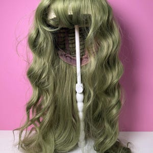 May include: A sage green wig with long, wavy curls and a blunt fringe. The wig is displayed on a white wig stand against a pink background. The wig's cap is visible, showing the construction.