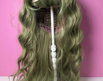 May include: A sage green wig with long, wavy curls and a blunt fringe. The wig is displayed on a white wig stand against a pink background. The wig's cap is visible, showing the construction.