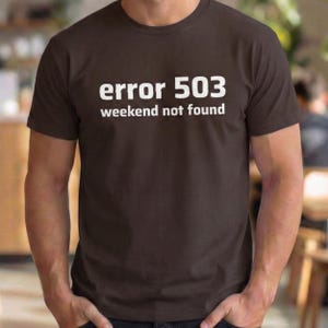 Könnte beinhalten: Ein braunes T-Shirt mit dem weißen Aufdruck "error 503 weekend not found". Das Shirt hat kurze Ärmel und einen Rundhalsausschnitt.
