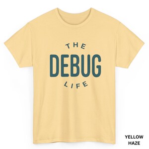 May include: Yellow haze t-shirt with the text "THE DEBUG LIFE" in teal. The t-shirt is a short-sleeved crew neck style. The text is centered on the front of the shirt.
