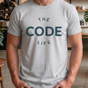 Op de afbeelding: Een lichtgrijs t-shirt met de tekst "THE CODE LIFE" in donkerblauwe letters. Het shirt is gemaakt van een zacht materiaal en heeft korte mouwen. De persoon draagt het shirt.