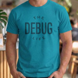 Può includere: T-shirt turchese con la scritta "THE DEBUG LIFE" in grigio scuro. La maglietta è a tinta unita e ha maniche corte. Il testo è centrato sul davanti della maglietta.