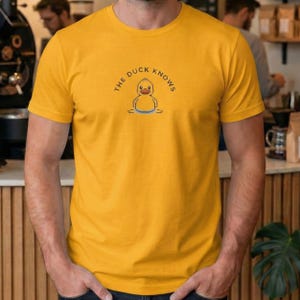 以下が含まれることがあります： マスタードイエローのTシャツ。ピクセル化されたアヒルのグラフィックと「THE DUCK KNOWS」のテキストが特徴です。デザインには、オレンジ色のくちばしと青い水線を持つ黄色いアヒルが含まれています。このTシャツは柔らかい素材で作られています。