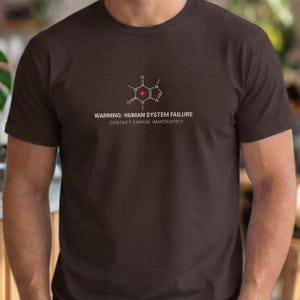 May include: A dark brown t-shirt featuring a caffeine molecule design in white and red. The text "WARNING: HUMAN SYSTEM FAILURE CONTACT COFFEE IMMEDIATELY" is printed below the graphic. The shirt is a crew neck.