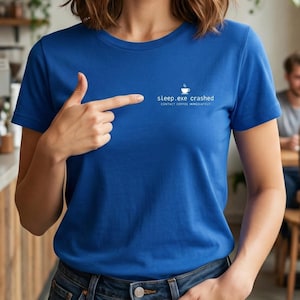 Può includere: Una maglietta blu reale con la scritta bianca "sleep.exe crashed CONTACT COFFEE IMMEDIATELY". La maglietta presenta una piccola grafica di tazza di caffè bianca sopra il testo. La persona indica il testo.