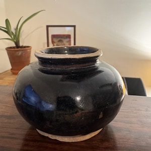 Peut inclure: Un pot en céramique rond, verni en noir, avec une large ouverture et un bord de couleur plus claire. Le pot repose sur une base de couleur claire et reflète la lumière ambiante. Une plante en pot et une œuvre encadrée sont visibles en arrière-plan.