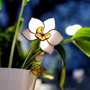 May include: A decorative stained glass flower in white and yellow, with a silver stem and leaves. It's set in a white pot with green foliage, against a blurred blue backdrop.