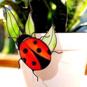 Puede incluir: Una mariquita de vidriera con alas rojas y negras y hojas verdes. La mariquita tiene puntos negros y cuelga de un alambre de metal. La mariquita está frente a una maceta blanca con una planta verde.