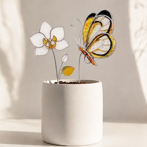 Può includere: Un vaso cilindrico in ceramica bianca contenente due ornamenti in vetro colorato su sottili steli metallici. Un ornamento è un fiore di orchidea bianca con il centro giallo, l'altro è una farfalla gialla e nera con ali traslucide.