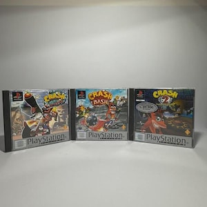 Può includere: Tre custodie di videogiochi per PlayStation con Crash Bandicoot. Le custodie sono nere con illustrazioni colorate. I titoli includono "Crash Bandicoot", "Crash Bash" e "Crash Bandicoot 2". Il logo PlayStation è visibile su ogni custodia.