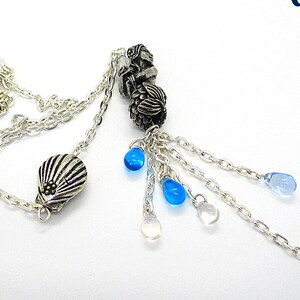 Puede incluir: Un collar de cadena de plata con un dije de concha y cuentas de vidrio azul. El collar tiene un borla de cadenas más pequeñas con cuentas azules en el extremo.