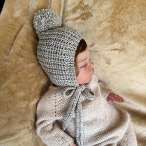Peut inclure: Un bonnet crocheté gris clair avec un pompon assorti sur le dessus et de longs liens au menton, associé à une grenouillère tricotée beige clair à manches longues. Le bonnet présente un motif texturé, tandis que la grenouillère est lisse.