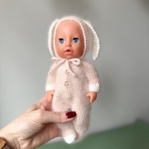 May include: A doll wearing a light pink knitted bunny outfit with a matching hood and button closure. The doll has blue eyes and a neutral expression. The outfit has white cuffs and a white bunny ear hood.