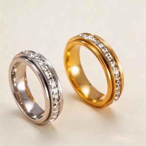 May include: Two rings, one silver-toned and one gold-toned, are displayed. Both rings feature a band of clear, sparkling stones. The silver ring has a double band design, while the gold ring has a single band.
