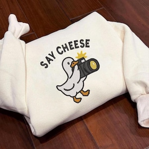 May include: Cream-colored sweatshirt with the phrase "SAY CHEESE" above an embroidered cartoon duck holding a camera. The duck is white with orange feet and beak. The camera is black and gold.
