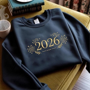 May include: A navy blue sweatshirt featuring the gold embroidered year "2026", with firework and floral designs. The sweatshirt is made of a soft material, suitable for casual wear. The design is centered on the chest.