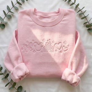 Pode incluir: Camisola rosa claro com desenhos bordados de um laço, a cabeça do Mickey, um castelo e corações. As mangas compridas são dobradas. A camisola é feita de um material macio e é adequada para uso casual.