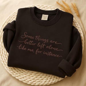 Puede incluir: Una sudadera negra con la cita "Some things are better left alone like me, for instance" en fuente cursiva. La sudadera está doblada sobre un salvamanteles circular tejido. Espigas de trigo secas en el fondo.