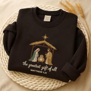 May include: Black sweatshirt featuring an embroidered Nativity scene with Mary, Joseph, and baby Jesus under a star. The text "the greatest gift of all" and "MATTHEW 1:21" are below the image. The sweatshirt is displayed on a woven mat.