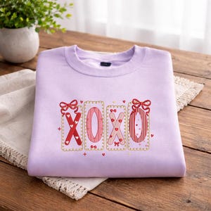 以下が含まれることがあります： 刺繍された「XOXO」のデザインが施された、薄紫色のスウェットシャツ。 赤とピンクの文字にハートとリボンのアクセント、金色のフレームが特徴です。 スウェットシャツは木製の表面に折りたたまれています。