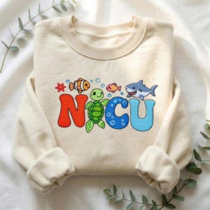 May include: Cream-colored sweatshirt with the word "NICU" in colorful embroidered letters. The letters are decorated with sea creatures, including a shark, turtle, and fish. The sweatshirt has long sleeves and a crew neck.