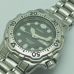 Peut inclure: Montre Casio argentée avec cadran noir. La montre dispose d'une lunette rotative avec des marqueurs de minutes, des aiguilles lumineuses et un affichage jour/date. Résistante à l'eau jusqu'à 20 bars.