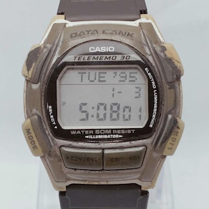 Może przedstawiać: Zegarek cyfrowy Casio Data Bank Telememo 30 w stylu vintage. Zegarek ma czarny pasek i zwietrzałą, beżową obudowę. Wyświetlacz cyfrowy pokazuje datę i godzinę. Zegarek posiada przyciski do trybu, wyboru, światła, wstecz i do przodu. Mierzy około 4 cm.