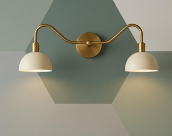 May include: A brass-toned wall sconce with two off-white, dome-shaped shades. The fixture has a curved arm design and a circular base. The sconce is mounted on a wall with a geometric pattern in shades of green and gray.