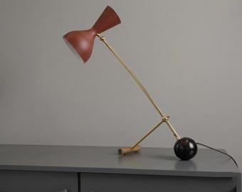 May include: A mid-century modern desk lamp with a red shade and brass arm. The lamp has a wooden base and a black spherical counterweight. The shade is perforated and angled, and the lamp is plugged into an electrical outlet.