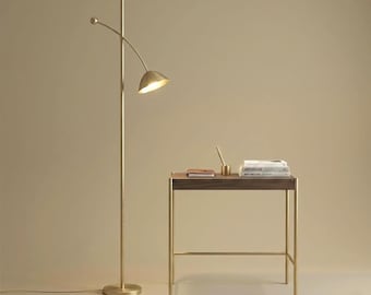 May include: A tall brass floor lamp with a single curved arm and a dome-shaped shade, positioned next to a rectangular wooden desk with a metal frame. The desk holds a small notebook, a pen in a holder, and a stack of three white books.