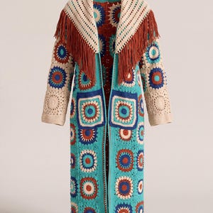 Può includere: Un lungo cardigan all'uncinetto aperto sul davanti con un vivace patchwork di motivi circolari e quadrati in turchese, crema, arancione ruggine e blu scuro. Il capo presenta un ampio collo a scialle color crema con lunghe frange color ruggine.