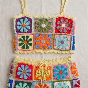 Op de afbeelding: Een tweedelige gehaakte set bestaande uit een mouwloze crop top en een rok. Beide kledingstukken zijn opgebouwd uit veelkleurige vierkante patches, elk met een verhoogd bloemmotief in diverse kleuren zoals rood, blauw, oranje en groen.