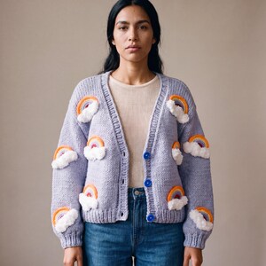 May include: A light purple knit cardigan featuring a V-neckline, long sleeves, and a button-front closure with three blue buttons. The garment is decorated with multiple embroidered rainbow motifs, each accented with a fluffy white cloud applique.