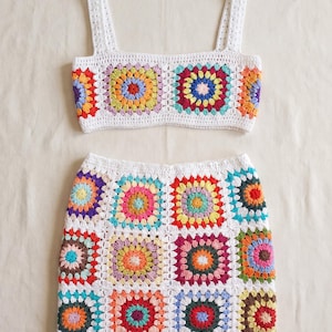 Può includere: Un completo in due pezzi all'uncinetto composto da un top corto e una minigonna. Entrambi i capi sono realizzati in filato bianco con motivi a piastrelle granny colorate che presentano cerchi concentrici nei toni del rosso, giallo, blu, verde e viola.