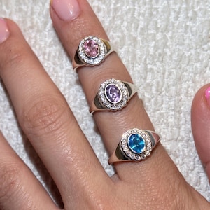 May include: Three silver rings featuring oval-cut centre stones in pink, purple, and blue. Each ring has a scalloped halo setting encrusted with small clear stones. The bands are smooth and polished, showcasing a classic design with vibrant gemstone colours.