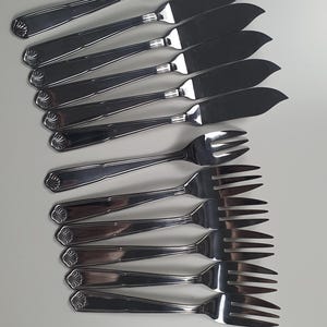 May include: A collection of stainless steel butter knives and forks. The knives feature a broad, rounded blade, and the forks have three prongs. Each piece of cutlery has a shell design on the handle.