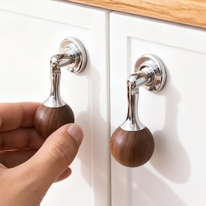 May include: Two cabinet door handles with a chrome and wood design. Each handle has a dark brown, spherical wooden knob and a curved chrome stem. One handle is being held by a hand. The cabinet doors are white.