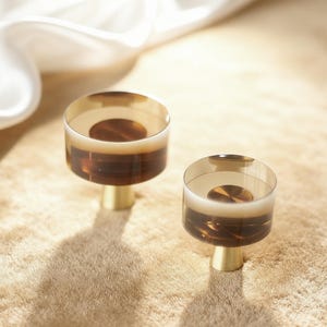 Könnte beinhalten: Zwei dekorative Schrankknöpfe. Jeder Knopf hat ein zylindrisches Design mit einem goldfarbenen Sockel und einer transparenten, bernsteinfarbenen Oberseite. Sie sind vor einem neutralen, strukturierten Hintergrund mit weicher Beleuchtung platziert.