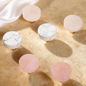 May include: A collection of decorative cabinet knobs. The knobs feature a round design with a variety of materials, including pink quartz and white marble with gray veining. Each knob has a gold-colored base.