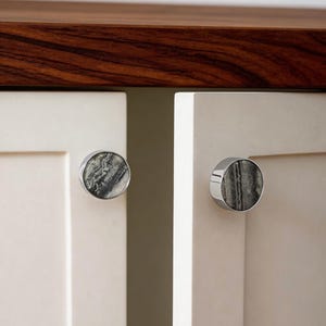 May include: Close-up of two cabinet knobs. Each knob features a circular, gray and white marbled stone set within a silver-colored metal frame. The knobs are mounted on off-white cabinet doors, with a dark wood countertop in the background.