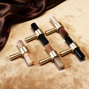 May include: Six cabinet knobs with brass fittings and cylindrical handles in various colours: black, brown, beige, and light wood grain. The knobs are arranged on a textured, golden-brown surface.