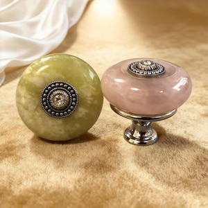 Könnte beinhalten: Zwei dekorative Schrankknöpfe. Einer ist ein hellgrüner, runder Knopf mit einer silberfarbenen, kreisförmigen Verzierung. Der andere ist ein rosa, runder Knopf mit einer silberfarbenen, kreisförmigen Verzierung und einem silberfarbenen Sockel.